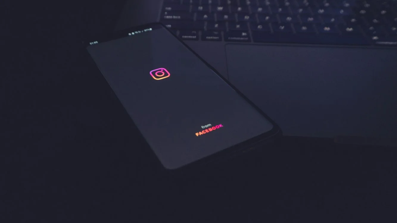 Celular em cima de um notebook mostra a primeira página do Instagram. A tela do celular e o ambiente estão escuros. A única luz vem do símbolo do Instagram.